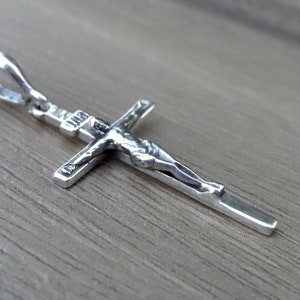 Puede incluir: Pendiente de plata en forma de cruz con una figura detallada de Jesucristo. El pendiente tiene un bucle en la parte superior para sujetarlo a una cadena.