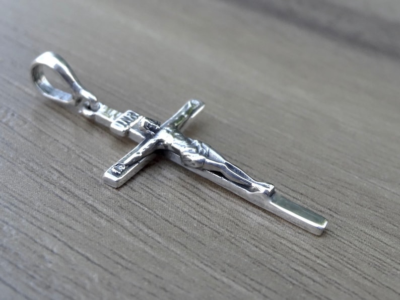 Puede incluir: Pendiente de plata en forma de cruz con una figura detallada de Jesucristo. El pendiente tiene un bucle en la parte superior para sujetarlo a una cadena.