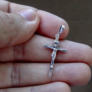 Puede incluir: Pendiente de plata en forma de cruz con una figura detallada de Jesucristo. El pendiente se sostiene en una mano.