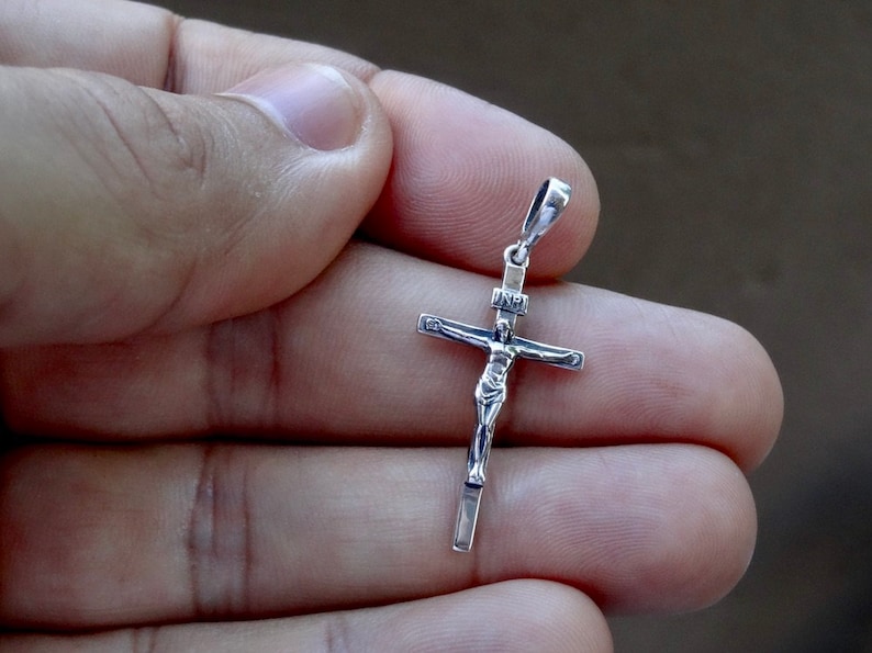 Puede incluir: Pendiente de plata en forma de cruz con una figura detallada de Jesucristo. El pendiente se sostiene en una mano.