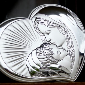 Puede incluir: Una placa de plata en forma de corazón con una imagen en relieve de la Virgen María sosteniendo a un bebé Jesús. La placa tiene un fondo texturizado con líneas radiantes.
