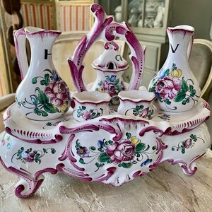 Könnte beinhalten: Ein weißes Keramik-Waschtisch-Set mit floralen Designs und lila Akzenten. Das Set enthält zwei Krüge mit den Buchstaben "H" und "V", zwei kleine Behälter und ein zentrales Stück mit Deckel, alles auf einem dekorativen Tablett.