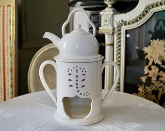 May include: White ceramic teapot with a built-in warmer. The warmer has a cut-out design and a space for a tea light candle. The teapot has a lid and a handle.