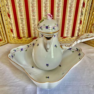 Pode incluir: Uma chocolateira de porcelana branca com bandeja festonada a condizer, decorada com flores azuis pintadas à mão, vinhas verdes e bordas douradas. A tampa é rematada por um botão de rosa esculpido e o bule tem uma pega longa e ornamentada.