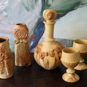 Puede incluir: Un conjunto de cuatro piezas de cerámica en color marrón claro con diseños intrincados. El conjunto incluye una decantador con un tapón único, dos tazas pequeñas y un jarrón alto.