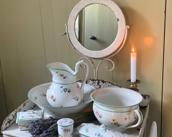 May include: A white porcelain bathroom set with floral designs. The set includes a pitcher, a bowl, a chamber pot, a soap dish, and a small jar. The items are arranged on a white painted wooden table with a round mirror and a lit candle.