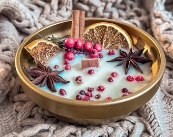 May include: A white candle with a gold rim in a gold bowl. The candle is decorated with dried orange slices, star anise, cinnamon sticks, and red peppercorns.