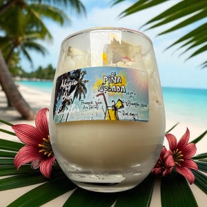 Könnte beinhalten: Eine klare Glaskerze mit weißem Wachs im Inneren, beschriftet mit "Piña Colada". Das Etikett zeigt eine tropische Getränkeillustration und die Worte "Magic Hour Candles". Rote Blumen und grüne Blätter umrahmen die Kerze, mit einer Strandszene im Hintergrund.