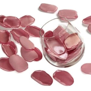 May include: A glass bowl filled with rose petals, surrounded by more rose petals on a white background. The petals are a soft pink color and have a satin finish.