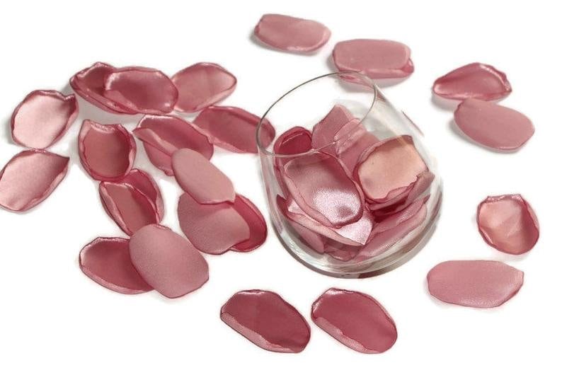 May include: A glass bowl filled with rose petals, surrounded by more rose petals on a white background. The petals are a soft pink color and have a satin finish.