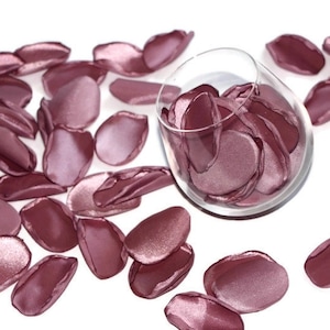 May include: A glass bowl filled with rose petals, with additional petals scattered around the bowl on a white surface. The petals are a soft, dusty rose color.