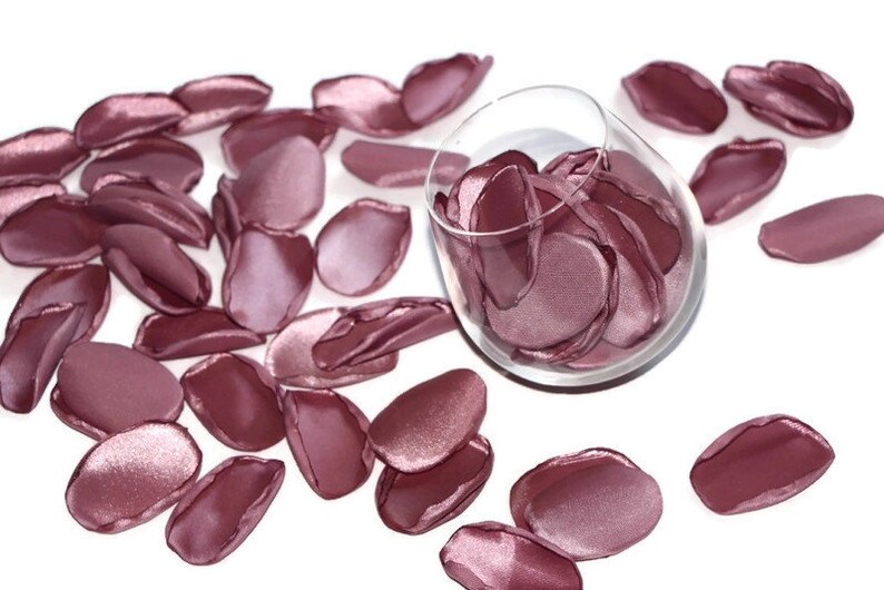 May include: A glass bowl filled with rose petals, with additional petals scattered around the bowl on a white surface. The petals are a soft, dusty rose color.