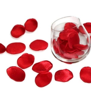May include: Red satin rose petals scattered on a white surface, with a glass vase filled with more petals.