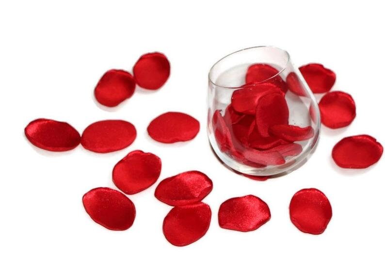 May include: Red satin rose petals scattered on a white surface, with a glass vase filled with more petals.