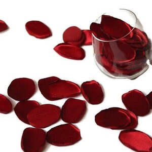 May include: Red satin rose petals scattered on a white surface. Some petals are in a clear glass vase.