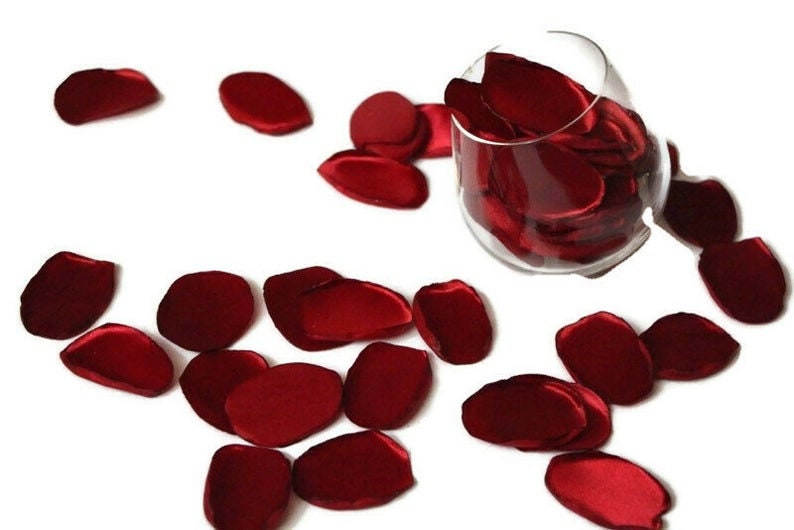 May include: Red satin rose petals scattered on a white surface. Some petals are in a clear glass vase.