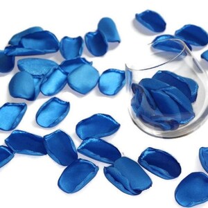 May include: A scattering of blue satin rose petals on a white surface. Some petals are in a clear glass vase.
