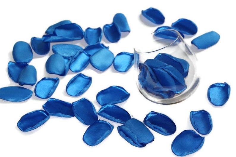 May include: A scattering of blue satin rose petals on a white surface. Some petals are in a clear glass vase.