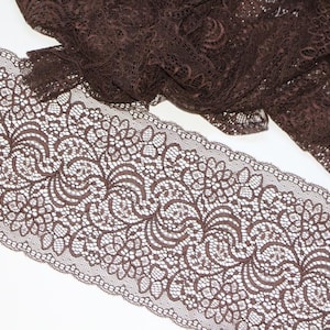 May include: A wide strip of dark brown lace fabric featuring an intricate floral and scalloped edge pattern. The lace is semi-transparent with a detailed, repeating design that runs horizontally across the length of the fabric trim.