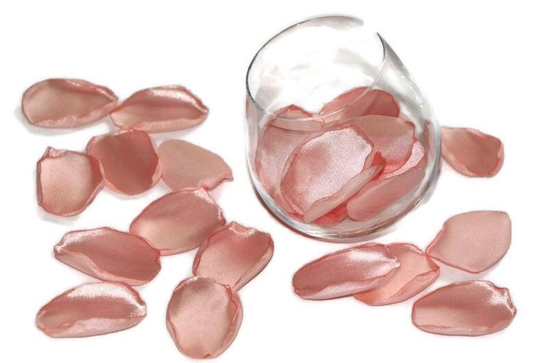 May include: A scattering of blush pink satin rose petals, some in a clear glass vase, others on a white surface.