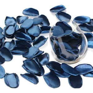 May include: A glass bowl filled with blue satin rose petals, with additional petals scattered around the bowl on a white background.