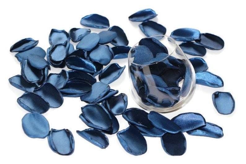 May include: A glass bowl filled with blue satin rose petals, with additional petals scattered around the bowl on a white background.