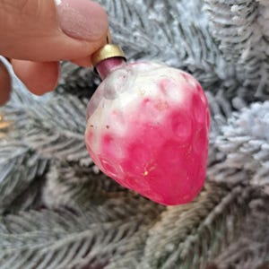 May include: A vintage Christmas ornament shaped like a strawberry. The ornament is primarily pink with white accents and a gold cap. The strawberry-shaped decoration is held against a backdrop of frosted evergreen branches.