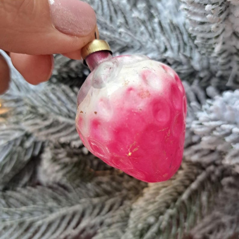 May include: A vintage Christmas ornament shaped like a strawberry. The ornament is primarily pink with white accents and a gold cap. The strawberry-shaped decoration is held against a backdrop of frosted evergreen branches.