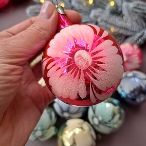 May include: A pink glass Christmas ornament with a white floral design. The ornament is round with a silver metal cap and a textured, frosted appearance. Other ornaments in various colors are visible in the background.