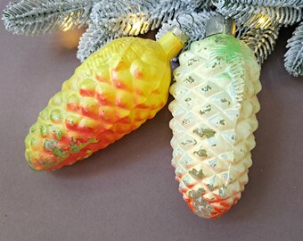 May include: Two vintage Christmas ornaments shaped like pine cones. One is yellow and red, the other is off-white and green. The ornaments are placed on a brown surface with a frosted evergreen branch in the background.