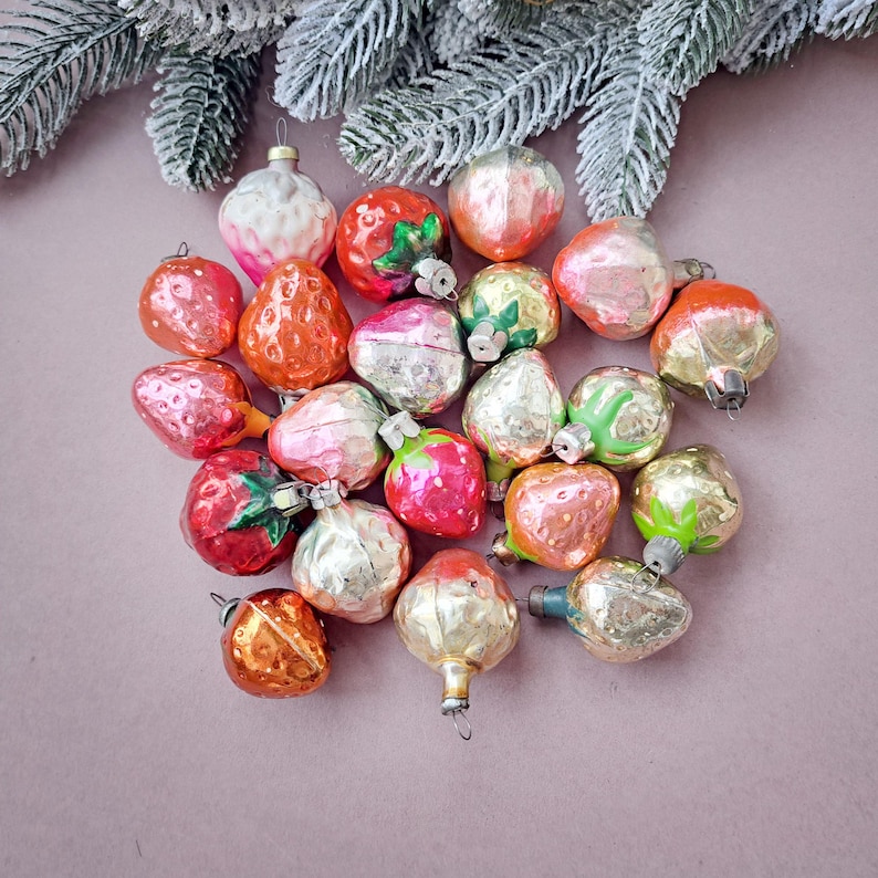 May include: A collection of vintage Christmas ornaments in the shape of fruits, including strawberries, apples, and pears. The ornaments are in various colors, such as red, orange, pink, and gold, with silver caps. A frosted evergreen branch is in the background.