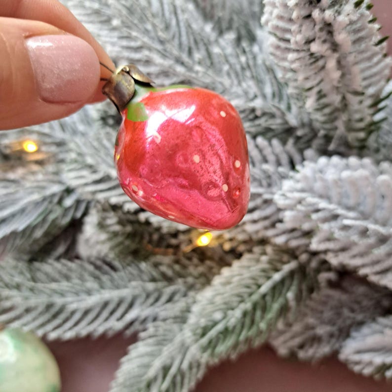 May include: A red strawberry Christmas ornament with white dots and a green stem. The ornament is held in front of a frosted Christmas tree. The ornament is a festive decoration.