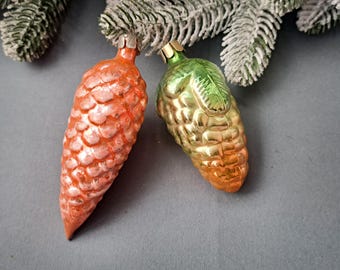 May include: Two vintage glass Christmas ornaments, one shaped like a pine cone in shades of orange and pink, and the other shaped like a bunch of grapes in shades of green and gold.