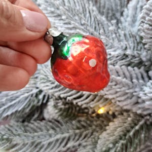 May include: A red glass strawberry Christmas ornament with a green stem and silver cap. The ornament has a glossy finish and a white dot. The ornament is held in front of a frosted Christmas tree.