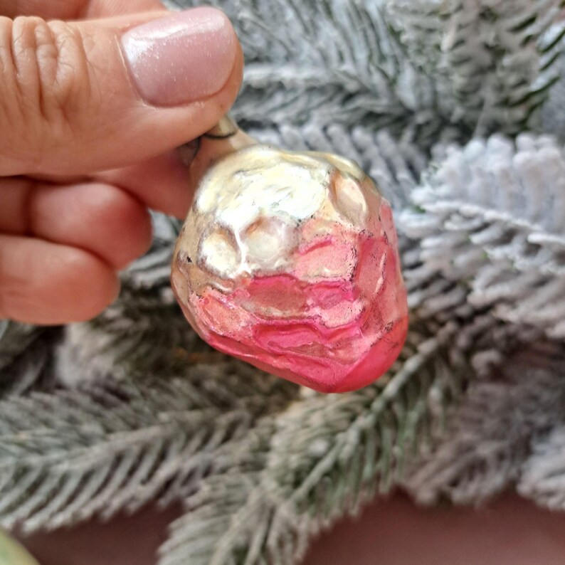 May include: A vintage Christmas ornament shaped like a strawberry. The ornament is primarily pink and white with a textured surface. The ornament is held in a hand, with a frosted evergreen branch in the background.