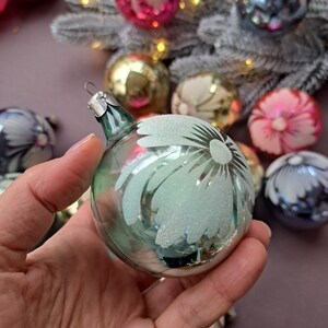 May include: A clear glass Christmas ornament with a frosted daisy design. The ornament is held in a hand, with other colorful ornaments and a frosted branch in the background. The ornament has a silver metal cap.