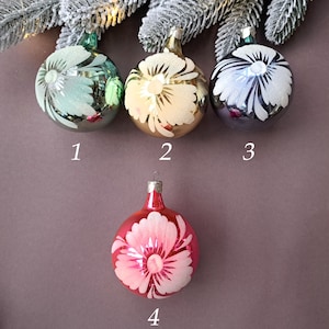 May include: Four glass Christmas ornaments. Three are round, in shades of green, gold, and silver, each with a white floral design. The fourth ornament is red with a pink and white floral design. All have metal caps.