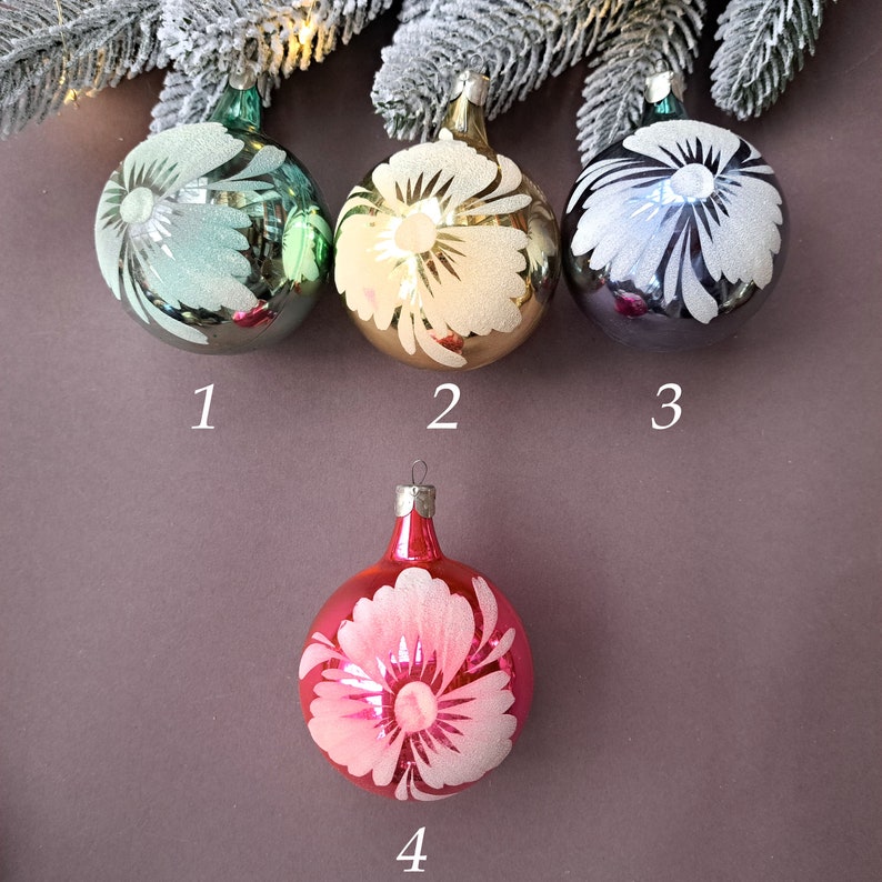 May include: Four glass Christmas ornaments. Three are round, in shades of green, gold, and silver, each with a white floral design. The fourth ornament is red with a pink and white floral design. All have metal caps.
