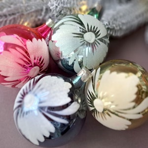 May include: Four glass Christmas ornaments in shades of pink, silver, and gold. Each ornament features a white floral design. The ornaments are round and have silver metal caps for hanging. The background includes a frosted white branch.