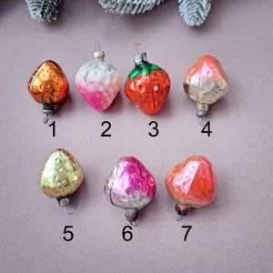May include: A collection of seven vintage glass Christmas ornaments in the shape of fruits. The ornaments are in various colors, including gold, pink, red, and green. Each ornament has a metal cap and a loop for hanging on a tree.