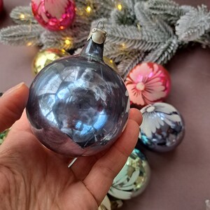 May include: A hand holds a vintage, iridescent blue Christmas ornament with a silver metal cap. Other colorful ornaments with floral designs and a frosted evergreen branch are in the background, suggesting a holiday theme.