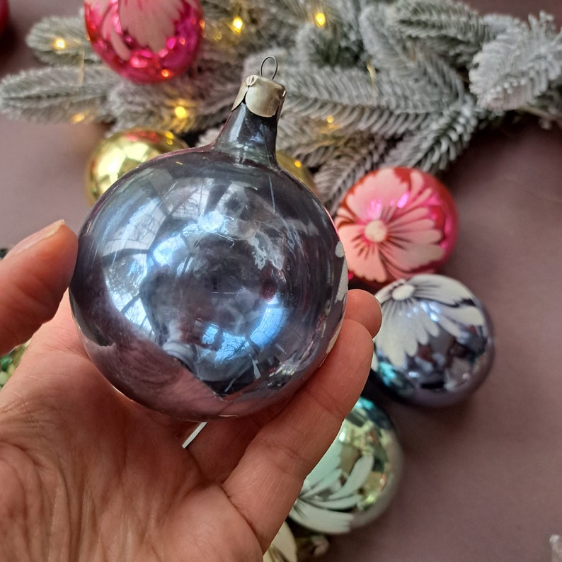 May include: A hand holds a vintage, iridescent blue Christmas ornament with a silver metal cap. Other colorful ornaments with floral designs and a frosted evergreen branch are in the background, suggesting a holiday theme.