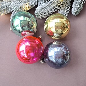 May include: Four vintage Christmas ornaments in teal, gold, red, and dark purple. Each glass ball has a silver cap and a reflective surface. The ornaments are arranged on a brown surface with frosted evergreen branches in the background.