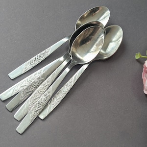 May include: Five silver spoons with floral and leaf patterns on the handles, arranged on a dark grey surface. The spoons are made of shiny metal, with pink and white flowers in the background. The spoons are a classic cutlery design.