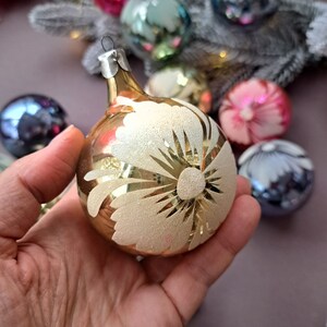 May include: A gold-colored Christmas ornament with a white floral design. The ornament is round and has a silver metal cap. Other colorful ornaments are visible in the background, along with a frosted branch.