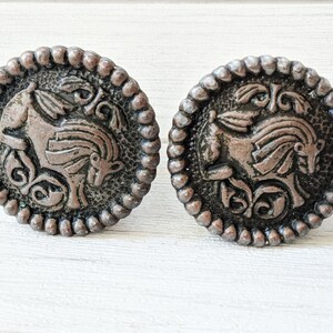 May include: Two antique bronze-colored cabinet knobs. Each knob features a detailed relief of a horse with flowing mane and decorative scrollwork. The knobs have a beaded edge and are set against a white background.