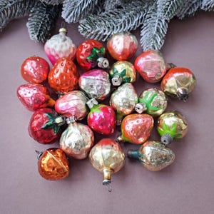 May include: A collection of vintage Christmas ornaments in the shape of strawberries and other fruits. The ornaments are in various colors, including red, pink, gold, and green. Some have silvered surfaces. A sprig of frosted evergreen branches is in the background.