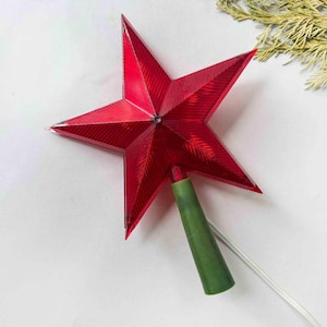 Puede incluir: Una estrella roja de cinco puntas para la parte superior del árbol con una superficie texturizada. La estrella tiene una base verde y un cable eléctrico blanco. Decoración navideña clásica.