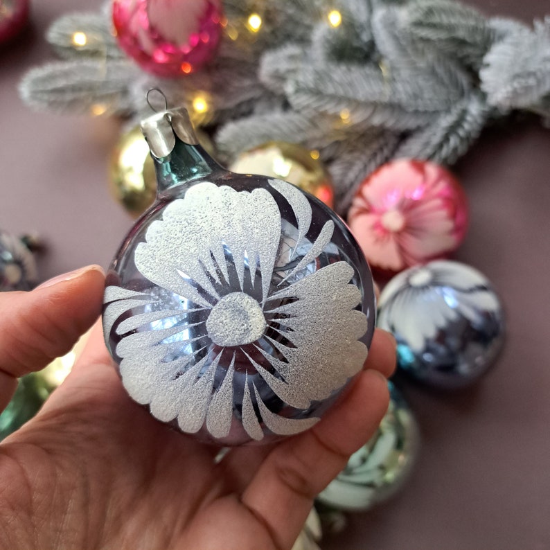 May include: A clear glass Christmas ornament with a silver floral design. The ornament is round and has a silver metal cap. Other colorful ornaments are in the background, along with a frosted pine branch.