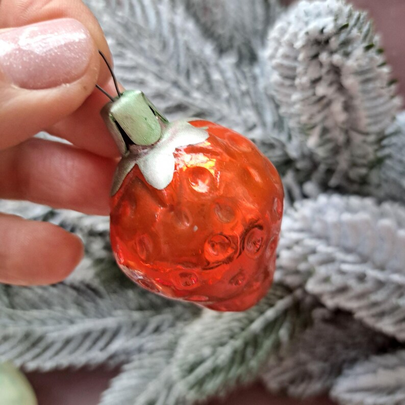 May include: A vintage Christmas ornament shaped like a strawberry. The ornament is a vibrant red with a textured surface, a green stem, and a white collar. It is held against a backdrop of frosted evergreen branches.